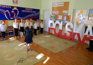 Lenka recytuje wiersz do mikrofonu, obok stoi duży napis biało -czerwonyPolska