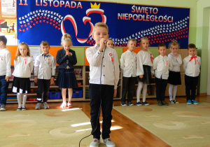 Łukasz recytuje wiersz do mikrofonu na tle dzieci z grupy oraz dekoracji