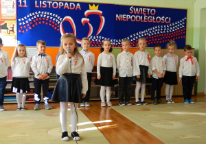 Amelka recytuje wiersz do mikrofonu na tle dzieci z grupy oraz dekoracji
