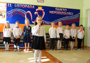 Gabrysia recytuje wiersz do mikrofonu na tle dzieci z grupy oraz dekoracji