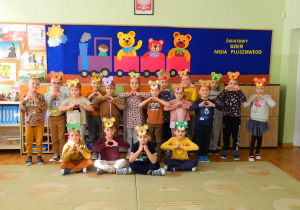 Dzieci z zerówki stoją na tle dekoracji o misiu w opaskach na głowach z misiem i pokazują z rąk kształt serca