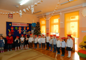 Dzieci stoją na tle dekoracji w czerwonych biretach czekając na wystep