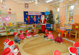 Dzieci z grupy Wiewiórek siedzą na dywanie, a Mikołaj stoi przy choince
