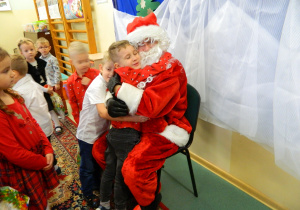 Mikołaj siedzi na krześle a dzieci przytulają się do niego