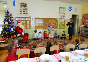 Dzieci siedzą na dywanie obok Mikołaja a pani dyrektor składa wszystkich życzenia