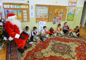 Dzieci z grupy Jeżyków siedzą na dywanie trzymając w rękch prezenty, obok Mikołaj pzry choince