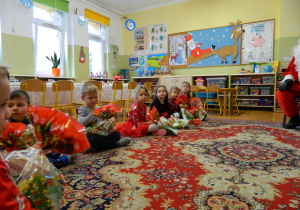 Chłopcy i dziewczynki siedza na dywanie i oglądaja prezenty od Mikołaja