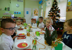 Dziewczynki i chłopcy z sześciolatków siedzą przy stole nakrytym białym obrusem i spożywają wspólnie obiad