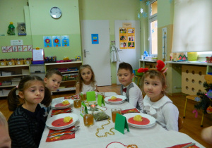 Dziewczynki i chłopcy z sześciolatków siedzą przy stole nakrytym białym obrusem i spożywają wspólnie obiad