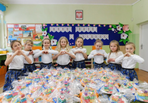 Dziewczynki z grupy Motylki stoją obok prezentów dla Dziadków, trzymają ręce ułożone w kształt serca