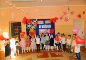 Dzieci z grupy Wiewiórek stoja na tle dekoracji z okazji Dnia Babci i Dziadka trzymając w rękach czerwone balony w kształcie serc