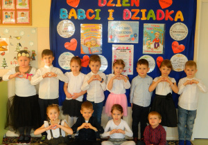 Dzieci z grupy Misiów stoją na tle dekoracji z okazji Dnia Babci i Dziadka , trzymają ręce ułożone w kształt serca