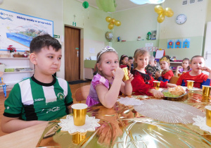 Dziewczynki i chłopcy z sześciolatków siedzą przy wspólnym stole spożywając owoce i ciasteczka oraz piją napoje podczas przerwy w tańcach