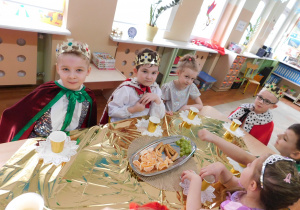 Dzieci w przebraniach siedzą przy stole nakrytym złotym obrusem, jedzą owoce, ciasteczka, piją napoje