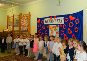 Dzieci z grupy Biedronek stoja w półkolu na tle dekoracji z czerwonych serc.