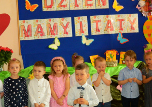 Chłopiec na tle dekoracji iinnych dzieci mówi wiersz do mikrofonu.