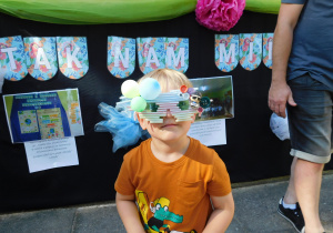 Chłopiec w okularach z małych baloników, guzików i słomek stoi na tle zdjęć z minionego roku przedszkolnego.