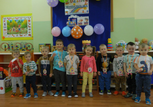 Maluchy z krasnali stoją na tle dekoracji z medalami na szyi z okazji Dnia Przedszkolaka.