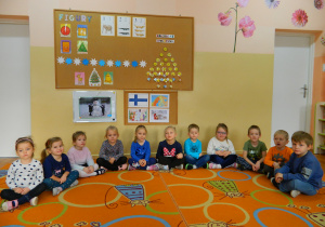 Przedszkolaki siedzą na dywanie w półkolu , w tle dekoracja z napisem Dzień Fiński