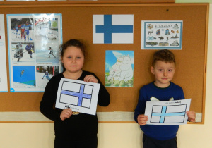 Dziewczynka i chłopiec stoją na tle tablicy z gazetką informacyjną ,trzymają w dłoniach narysowane flagi Finlandii
