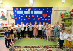 Dzieci z grupy Misiów stoją na tle dekoracji i pokazują z dłoni serduszko