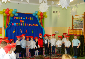 Przedszkolaki stoja na tle dekoracji w galowych strojach i czerwonych biretach na głowach