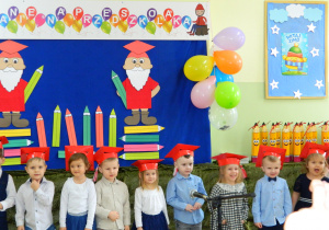 Przedszkolaki stoją na tle dekoracji z krasnali i kolorowych kredek