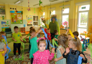 Pani Lucynka w przebraniu wiosny tańczy z dziećmi, które trzymają w dłoniach bibuły w kolorach wiosennych