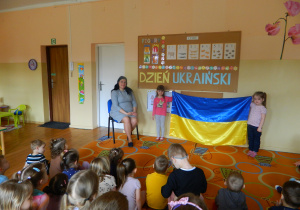 Dziewczynki trzymają dużą ukraińską flagę na tle tablicy , obok siedzi mama Anheliny a na przeciw dzieci