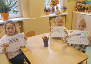 Dzieci siedza przystoliku i trzymają w rękach kolorowanki o dobrych słowach.