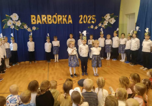 DZieci stoją na tle dekoracji i recytują wiersze do mikrofonu.