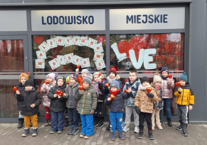 Dzieci stoją przed lodowiskiem miejskim trzymając w rękach czerwone serca.