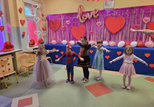 Przedszkolaki tańczą na tle czerwonych serc i balonów.