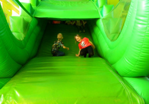 Dziewczynka i chłopak zjeżdżają na zielonej dmuchanej zjeżdżalni.