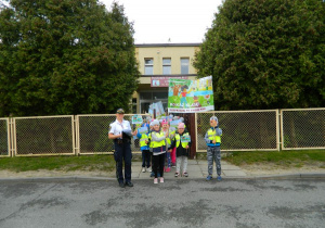 Dzieci wraz ze strażniczką miejską stoją przed wejściem do przedszkola z transparentem.