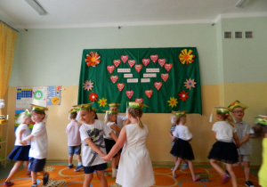 Dzieci z grupy Biedronek w biretach na głowach tańczą w parach na tle dekoracji.