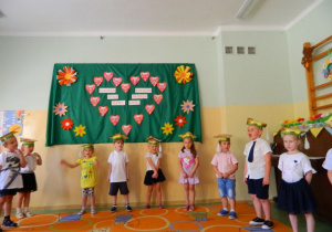 Dzieci z grupy Biedronek stoją na tle dekoracji i śpiewają piosenkę dla rodziców.