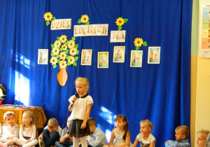 Dziewczynka recytuje wiersz na tle grupy i dekoracji.