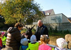 Pani Jola wraz z dziećmi głaszcze osiołka w ogrodzie kościelnym wraz z księdzem.