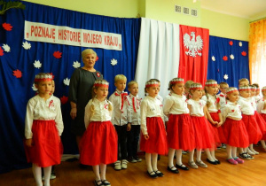 Pani dyrektor wraz z dziećmi na tle dekoracji śpiewa hymn narodowy.
