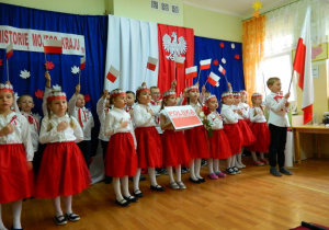 Dzieci tzrymaja w rękach flagi biało - czerwone oraz napis Polska Niepodległa i śpiewają.