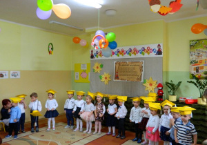 Dzieci z grupy Krasnali w żótych biretach na głowie stoją na tle dekoracji.