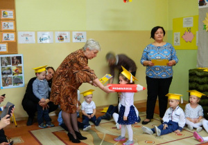 Na pierwszym planie Dyrektor pasuje na przedszkolaka przykładając czerwony duży ołówek na ramię dziecka.