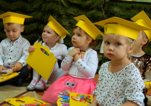 Dzieci siedzą na dywanie z czekoladkami i dyplomami w rączkach.