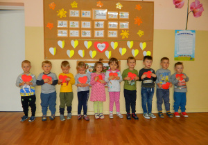 Dzieci z grupy Wiewiórek na tle dekoracji, trzymają czerwone serca w dłoniach