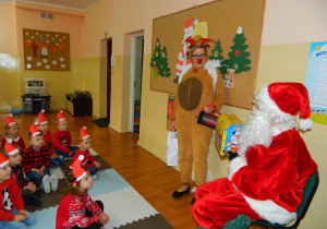 Mikołaj pokazuje dzieciom prezenty, obok stoi renifer