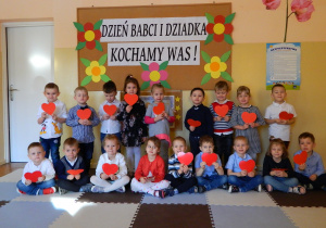Dzieci z grupy Wiewiórek stoją na tle dekoracji, trzymając w rączkach czerwone serca