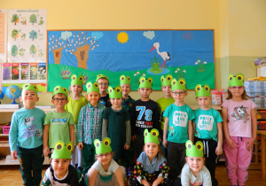 Dzieci z grupy Jeżyków stoją na tle dekoracji w opaskach żabek na głowach