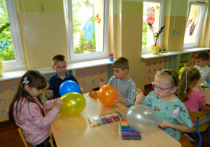 Dzieci z grupy Motylków siedzą przy stolikach i malują balony